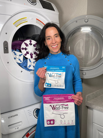 Wad-Free Inventor Cyndi Bray stands in a blue dress next to a washer and dryer holding a blue package of Wad-Free for Bed Sheets and an orchid package of Wad-Free for Blankets & Duvet Covers with both products showing in the open dryer