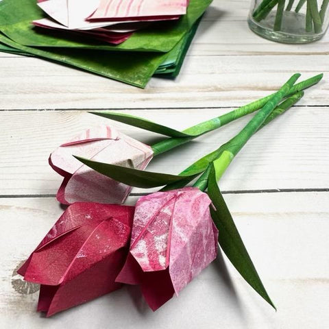 three red origami tulips with green stems lying on a table with a stack of green and red origami paper in the background