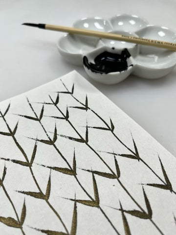 leaf-like gold ink marks on paper with a flower-shaped porcelain dish and brush in the background