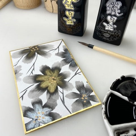 sumi flowers with metallic gold and silver accents on a shikishi board surrounded by ink bottles, a paintbrush and a porcelain palette filled with ink