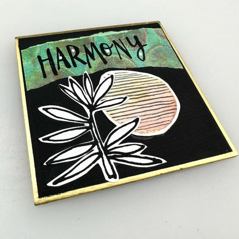 square shikishi board painted black and collaged with a cutout leaf and striped circle with the word "Harmony" across the top on a green banner