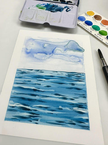 watercolor scene of ocean water and blue clouds with watercolor paints, swatch card and brush in the background