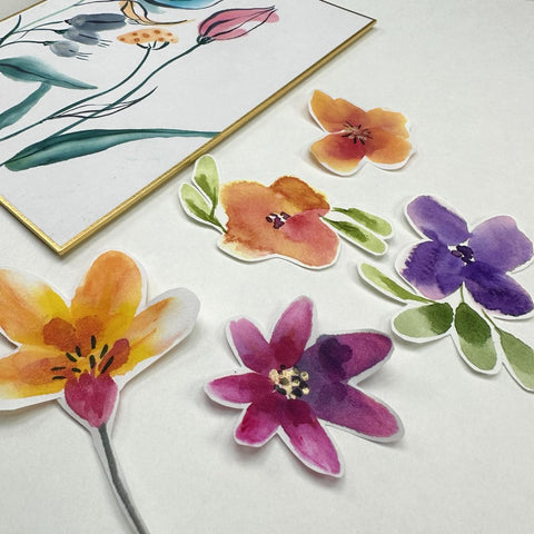 watercolor paper cut out flowers arranged on a table with a watercolor flower painting in the background