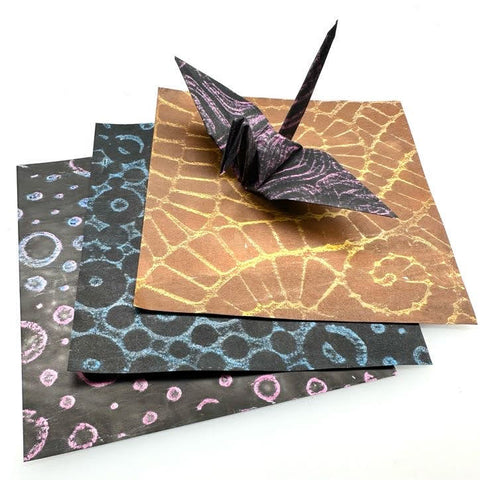 origami crane made of custom origami paper with crayon scribbles resting on top of three sheets of custom origami papers with crayon designs