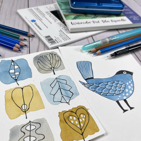 blue bird and pen drawn leaf doodles on top of watercolor squares with watercolor paper pads, watercolor pencils and waterbrushes around