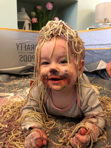 toddler lying in a crafty pod with colourful spaghetti on his head as he smiles at the camera