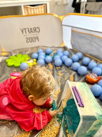 Child in red raincoat playing in a Crafty Pod with sensory items such as cereals, balls and slime