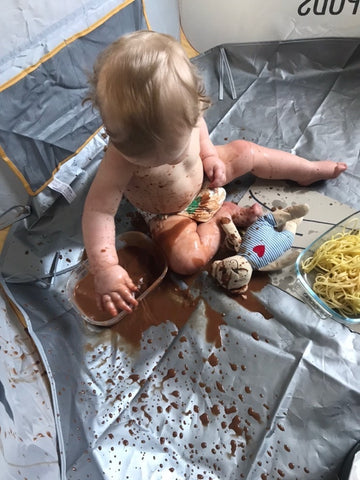 Toddler sat in Crafty Pot with a Messy Mat, playing with a container of thick brown liquid