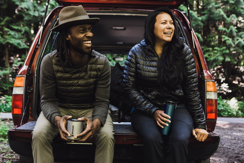 Man and woman sitting on the tailgate of a station wagon, each holding a Stanley titanium mug.