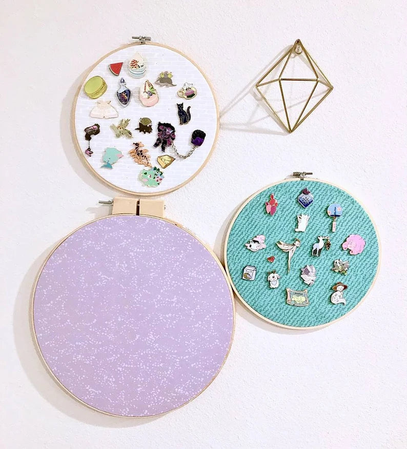 Counter clockwise, small embroidery hoop with white fabric and enamel pins, large embroidery hoop with lilac fabric and enamel pins, and a small embroidery hoop with teal fabric and enamel pins.