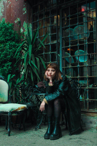 ALT TEXT: Woman In Emerald Green Trench Coat, Patterned Black Tights and Boots Sitting Hunched Over