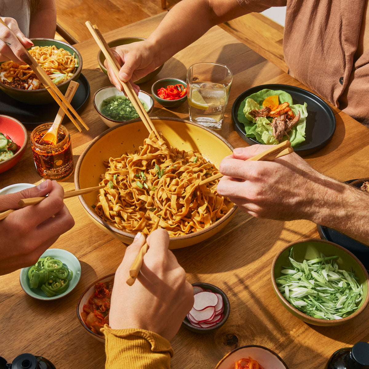 People sharing a meal of noodles and various side dishes, using chopsticks.