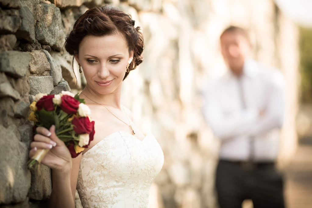A bride at a wall looking at her flowers with the groom in the back