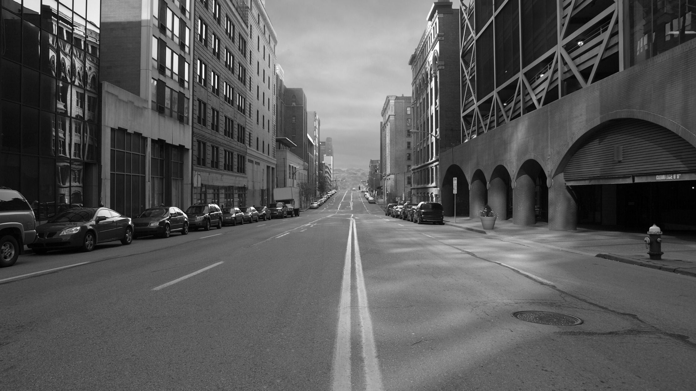 A black and white view of a long road with buildings on both sides and parked cars. 