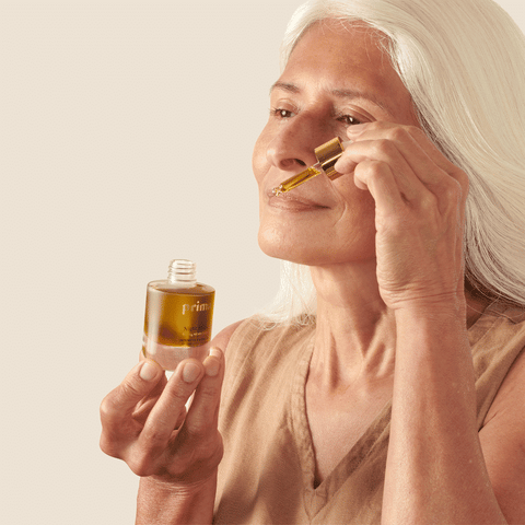 Edie Kahula Pereira is applying a face oil product (by Prima) into her skin . her medium length white hair is pulled away from her face. Edie is modeling this skincare product.