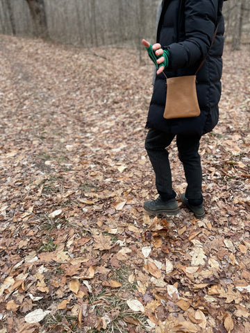 woman stands wearing x-small tan leather crossbody bag. its bottom corners are rounded. This bag is artisan made and handcrafted.