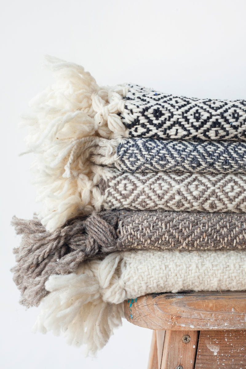 Wool throw blankets with diamond pattern weave in neutral tones and tassels stacked on top of a stool