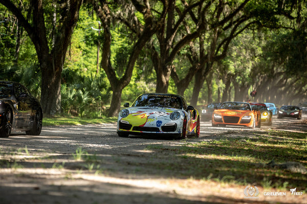 A group of cars participating in goldRush Rally drive under a canopy of trees