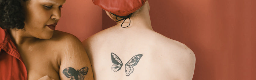 2 topless women, shoulder to shoulder, one facing front with butterfly tattoo on arm, the other facing back with butterfly tattoo on her back