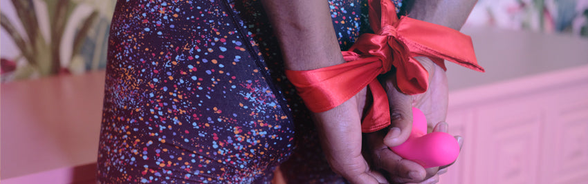 Closeup on a man's hand tied behind his back with a red ribbon, holding a Laya vibrator