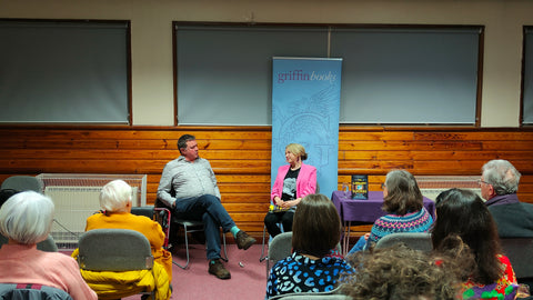 The image shows Fflur Dafydd and Jon Gower sat next to each other on chairs in front of a blue Griffin Books banner, in front of an audience of people. Fflur is smiling and they are in conversation.