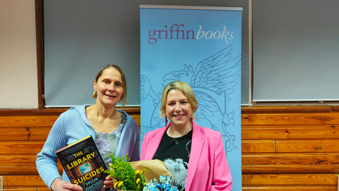 The photo shows Mel, the owner of Griffin Books, standing next to author Fflur Dafydd in front of a blue Griffin Books banner. They are both smiling and Fflur is holding a bunch of flowers. 