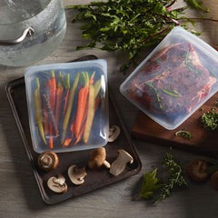 Two silicone stasher bags sit on a cutting board. One is filled with colorful carrots and the other contains marinating meat. 