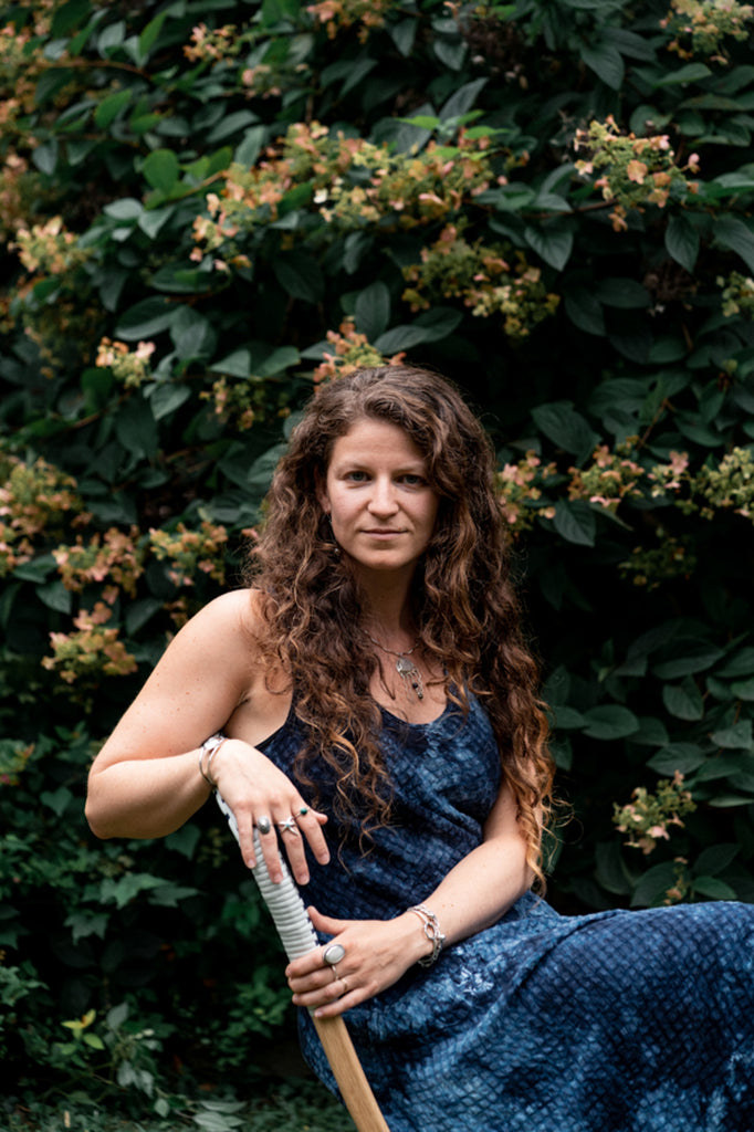 Hali MacLaren of HKM Jewelry portrait headshot sitting in chair in blue tie-dye dress with blooming greenery background photo by Traci Elaine 2023