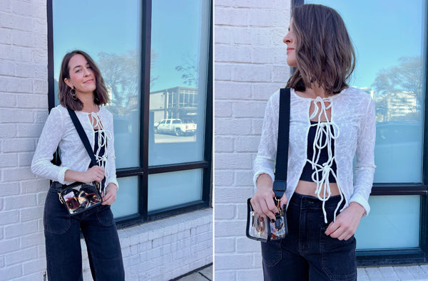 Haley is standing outside a storefront window. she is wearing a white cardigan and black denim and a clear handbag with a black strap. She is smiling because she loves her outfit.
