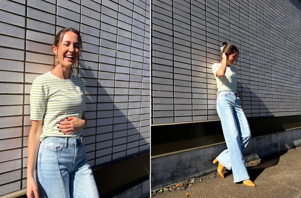 Haley is standing outside leaning against a white brick wall. She is wearing a striped white and green knit top, light wash denim and tan boots. She is smiling because she loves her outfit.