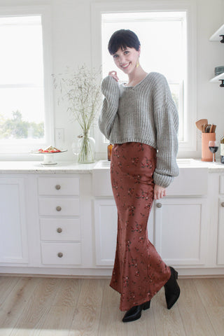 Tall person with pixi hair cut wearing a grey sweater and rust colored silk floral maxi skirt and black ankle boots. She is standing in a white kitchen.