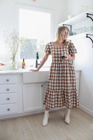 beautiful blonde girl standing in her white kitchen, getting ready to host a fall gathering. She is wearing a long gingham tiered a line dress with puff sleeves and white ankle boots. She is holding a glass of wine and waiting on her friends to arrive.