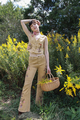 Model kennedy wearing the trinity cords and trinity vest holding a basket of goldenrod flowers. She is standing infront of a field full of goldenrod.