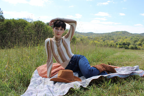 our model wearing a sweater, denim and boots, tipping her hat. she is sitting on a kantha quilt in a grassy field on a beautiful sunny day.