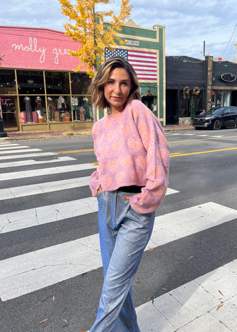 Lauren is standing outside our molly green hillsboro village location wearing a pink and purple knit sweater with hearts on it and silver glitter pants