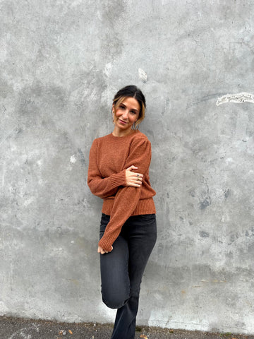 Lauren is standing in front of a grey concrete wall with an orange sweater and black denim pants