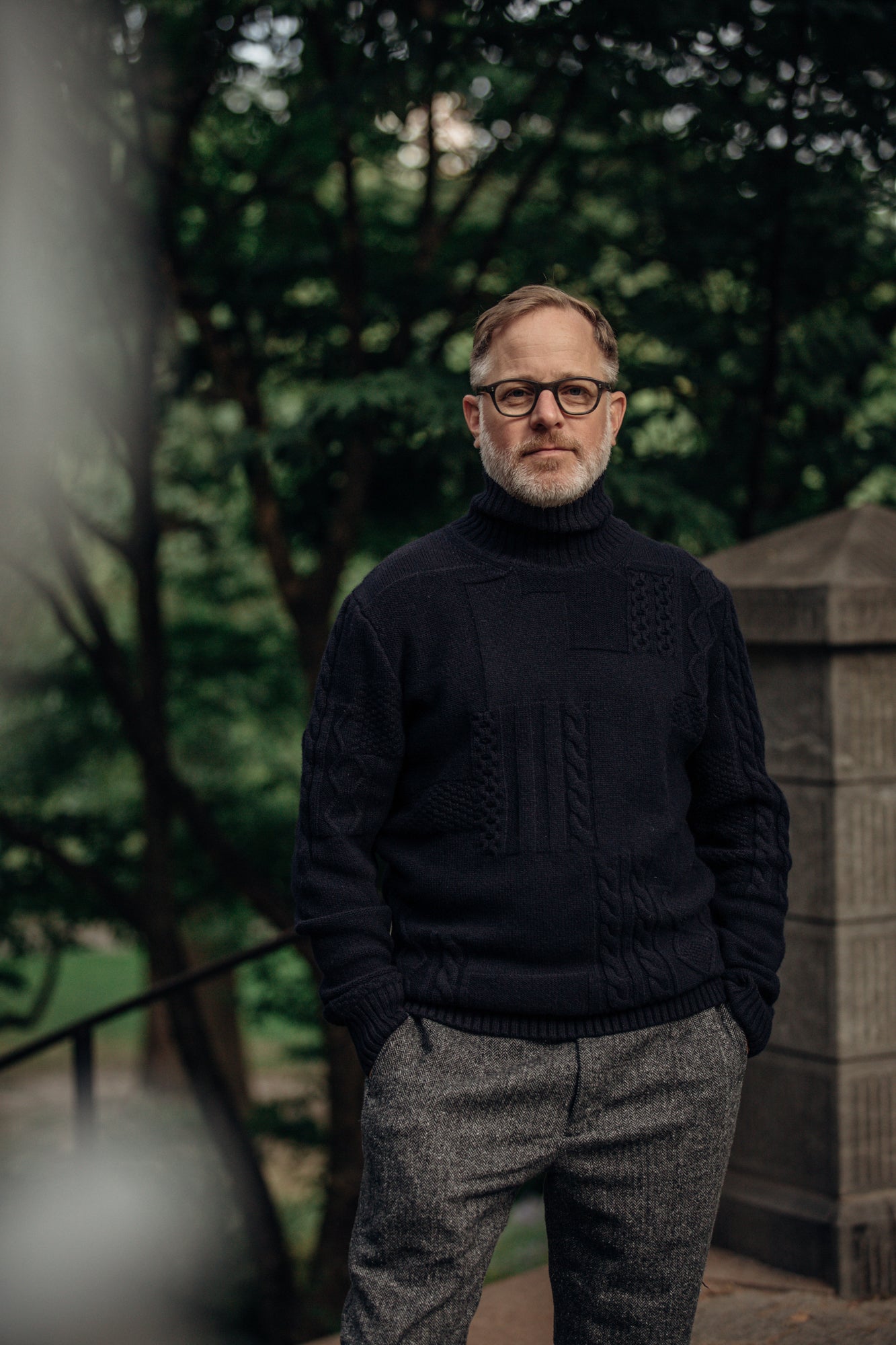 A man stands in a park wearing glasses and a black knit sweater.