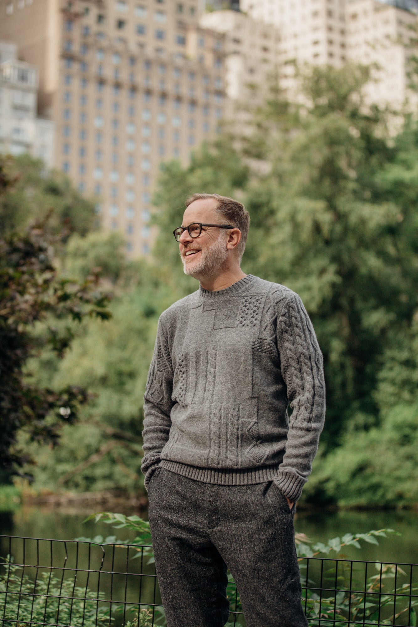 A man stands in a park wearing a glasses and a grey knit sweater.