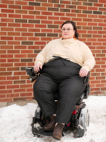 Val seated in Wheelchair in front of brick wall in Ramona trousers