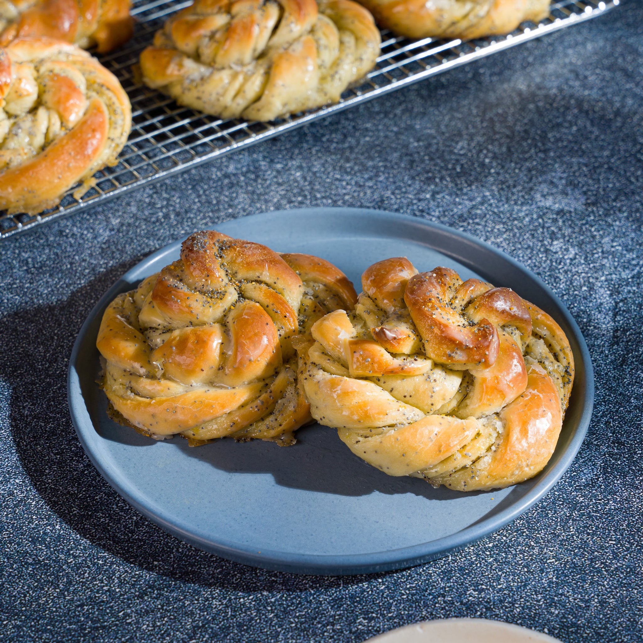 Two lemon poppy seed buns on a blue plate with more buns cooling on a rack above it