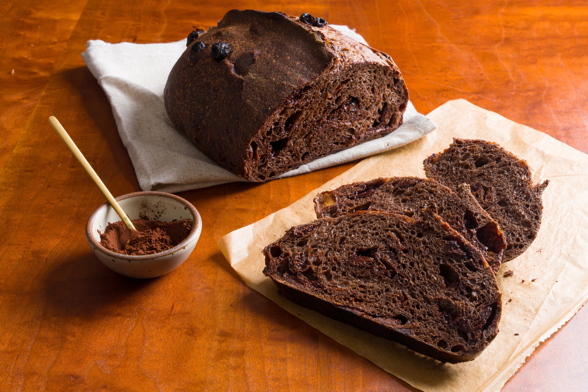 A loaf of chocolate cherry bread on a cream-colored cloth in the background  with cut slices on parchment paper in the foreground and a small ceramic bowl of cocoa powder to the left