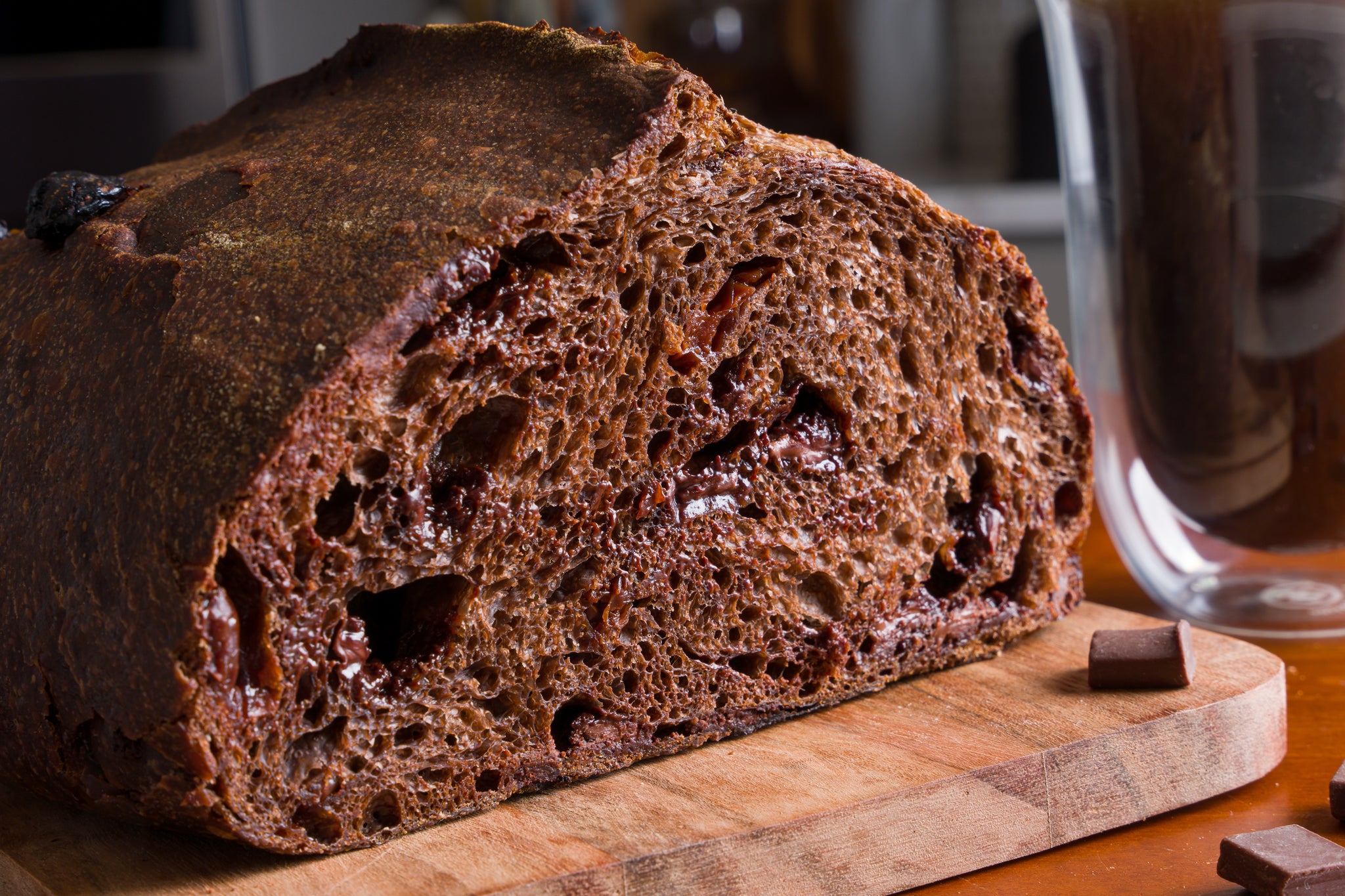 Close-up of the cut side of half of a loaf of chocolate cherry bread. Pockets of plump cherries and chunks of dark chocolate fill the cocoa-colored loaf