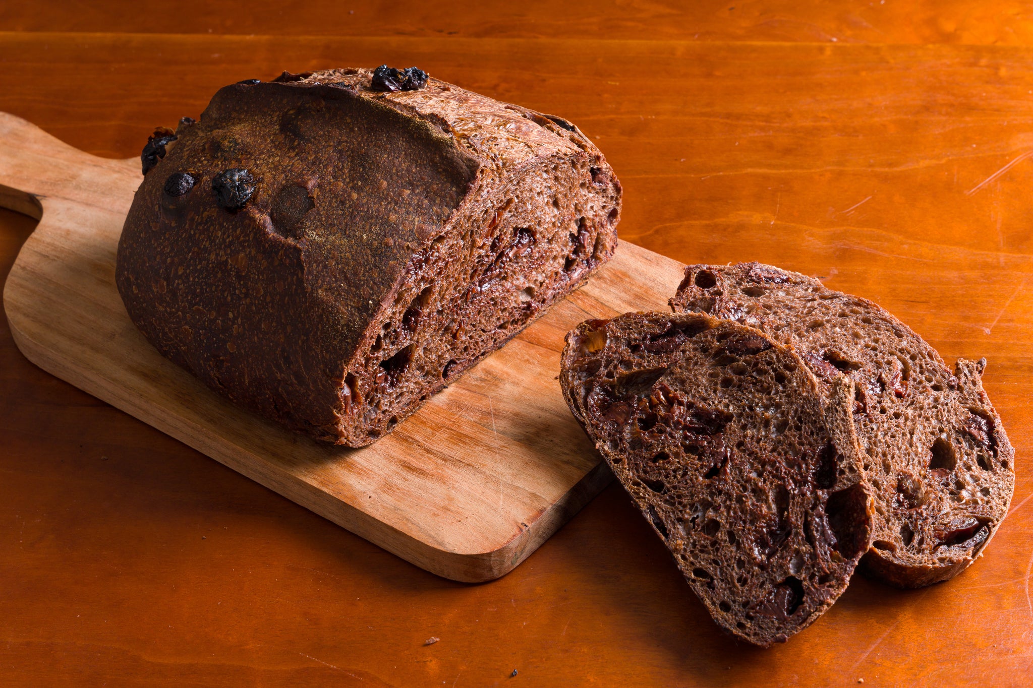 loaf of chocolate cherry bread on a wooden cutting board with two cut slices in front