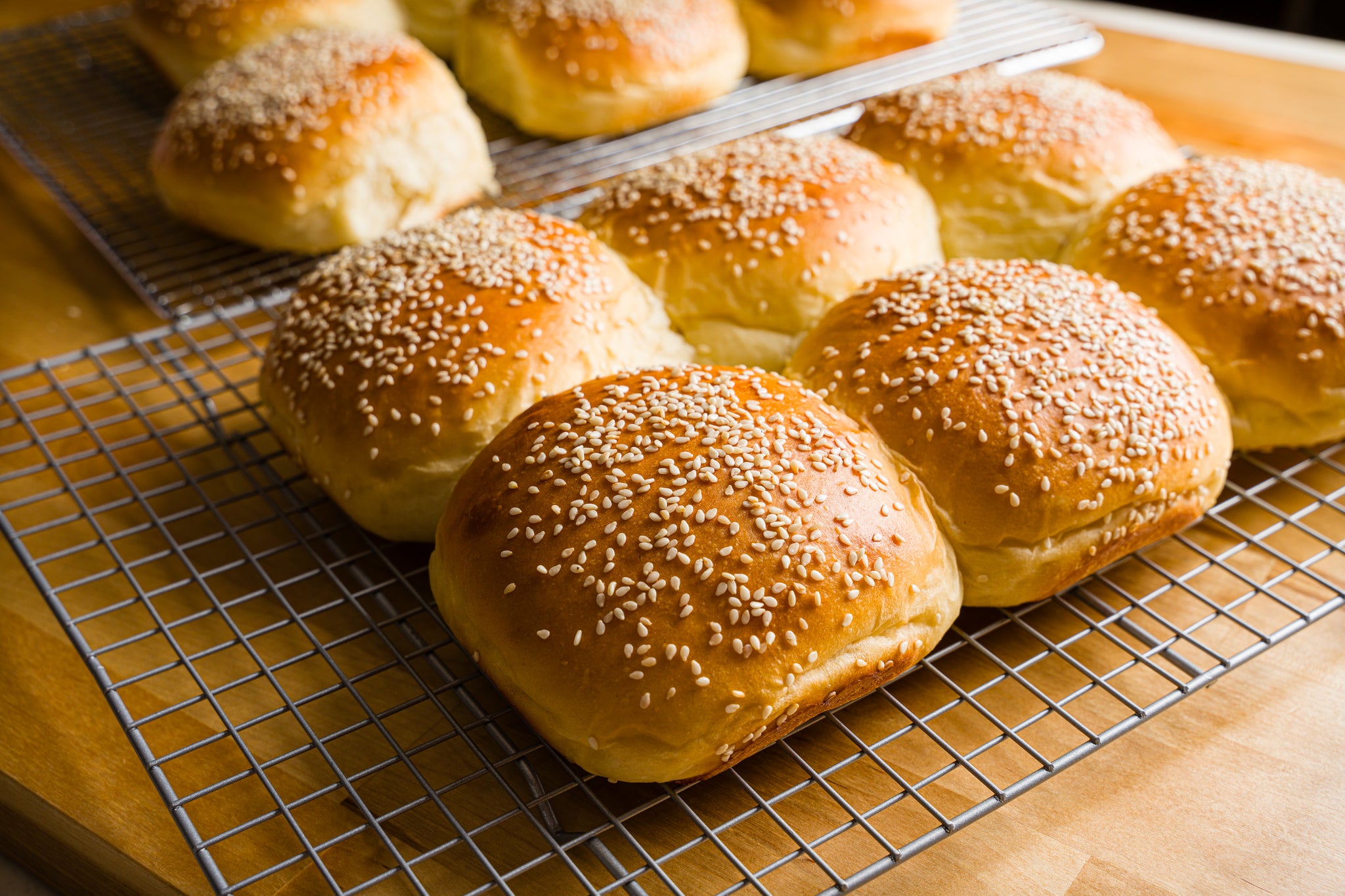 six golden brown burger buns with sesame seeds on top sit on a cooling rack