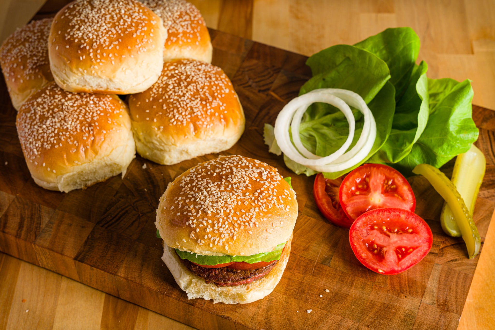 Burger buns on a cutting board next to lettuce, tomatoes, onion, and pickles. One assembled burger in the foreground.