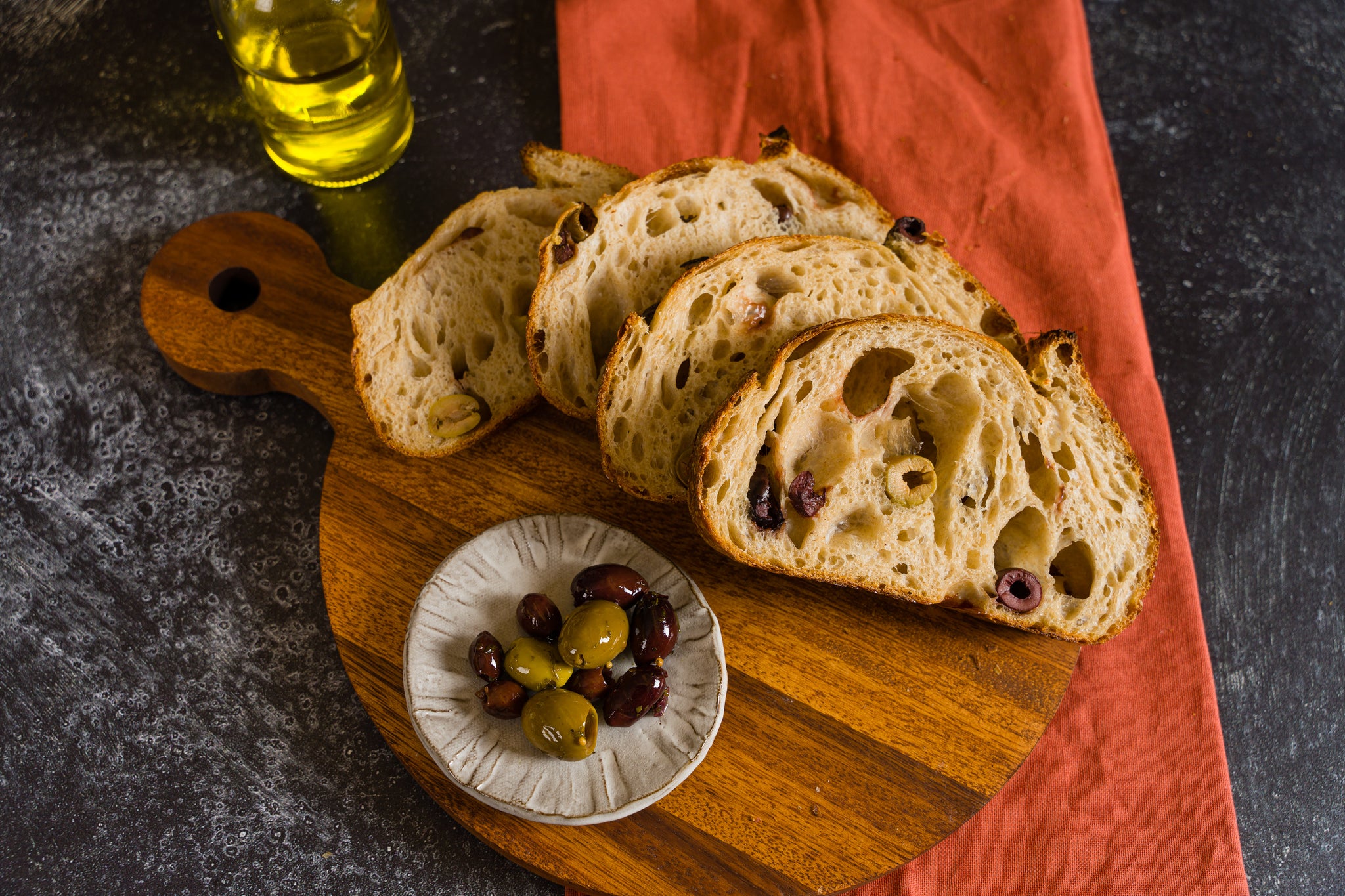 Slice of olive sourdough arranged on a round wooden cutting board with a dish of mixed olives to the left