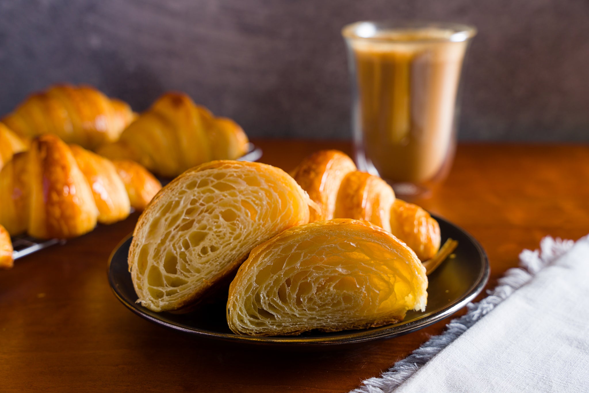 A croissant cut in half on a black plate with a rack of freshly baked croissants cooling in the background.