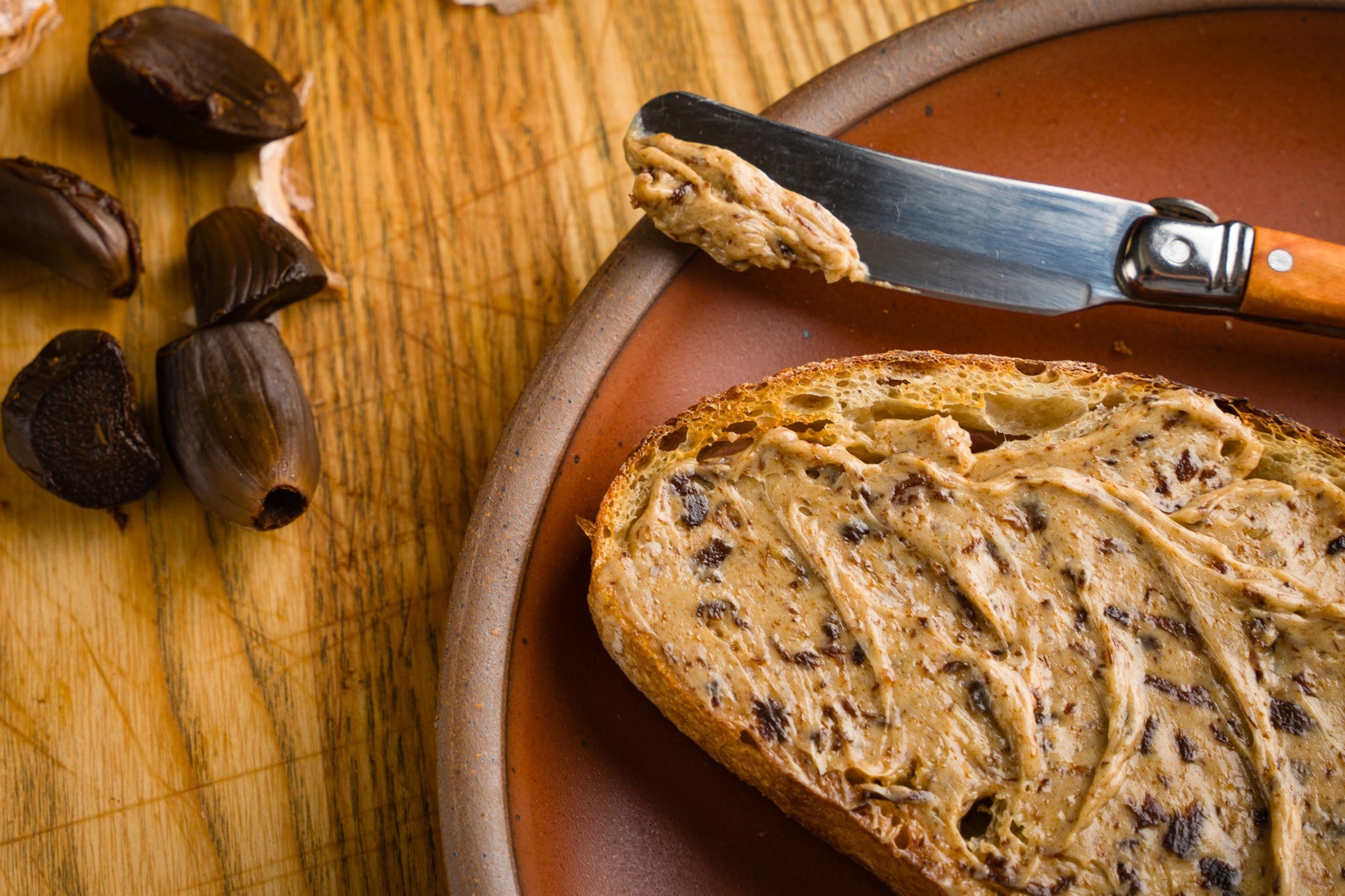 A slice of bread slathered in black garlic butter. Black garlic cloves sit off tho the left.