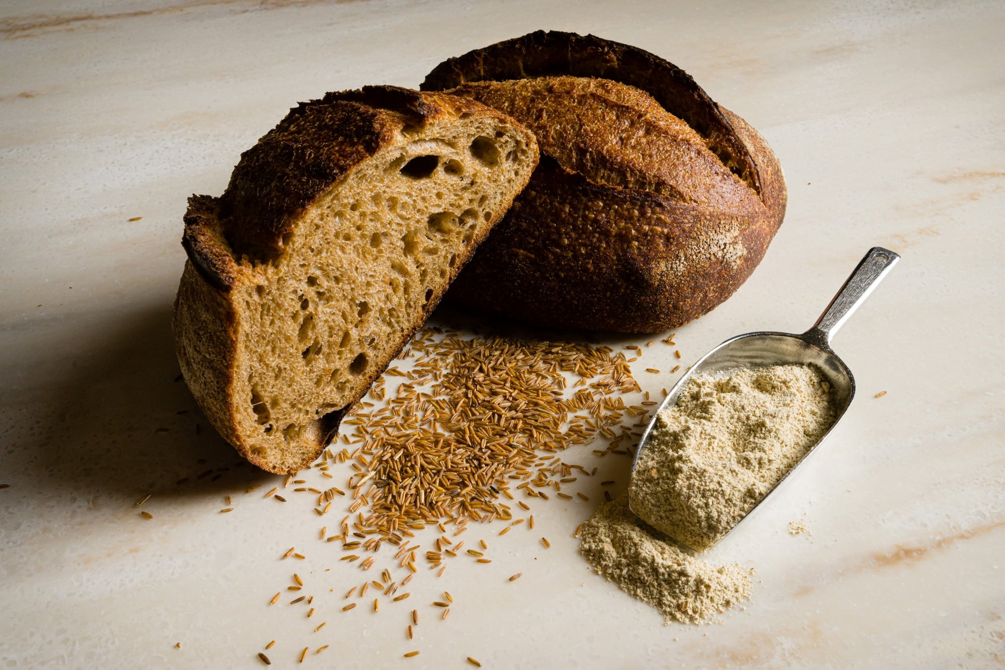 Kernza loaf cut in half with kernza grain scattered on the counter and a scoop of flour to the right