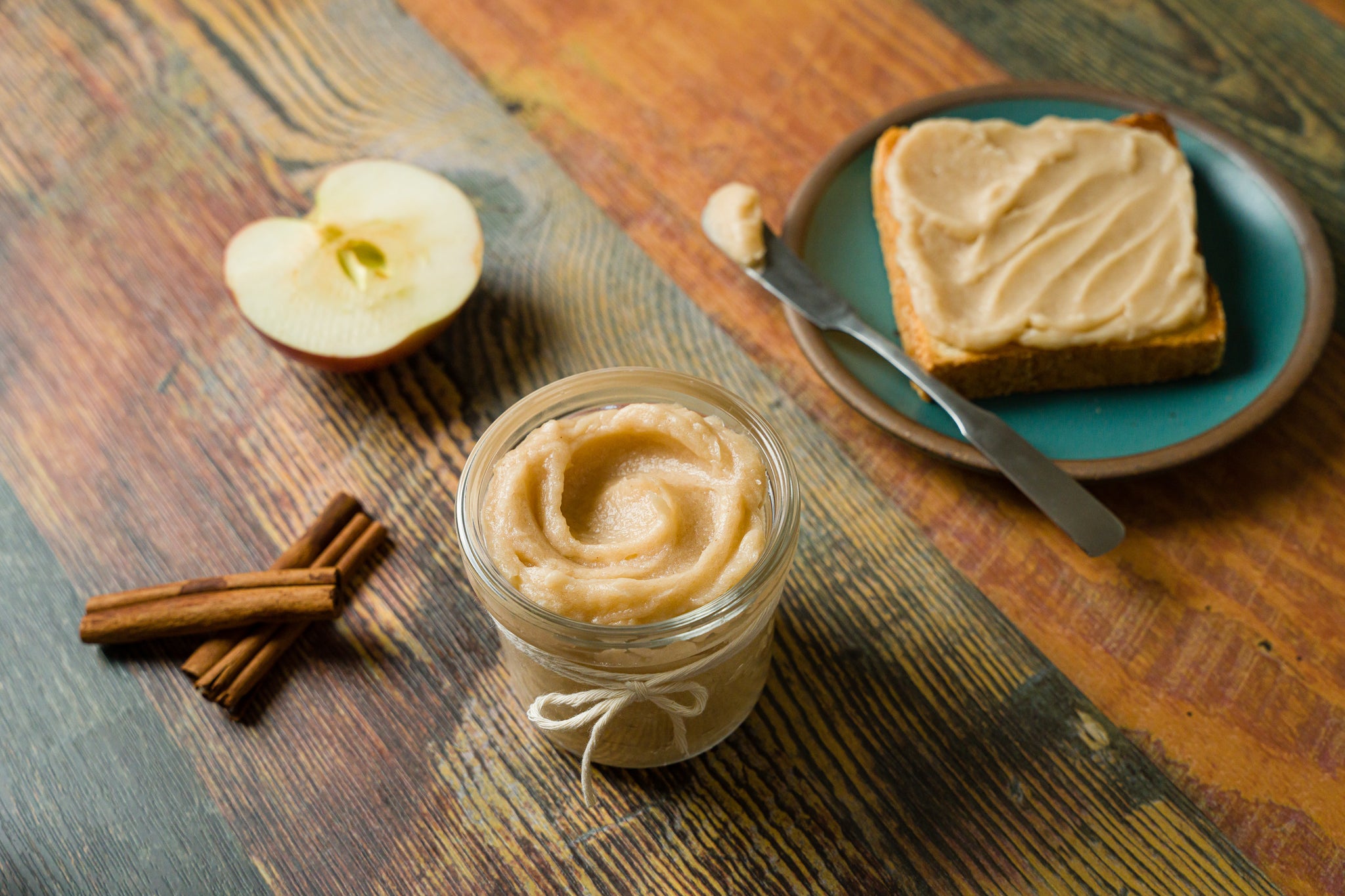 A glass jar filled with apple butter sits in the foreground with a slice of bread slathered in apple butter in the back right corner and half of a cut apple in the left corner